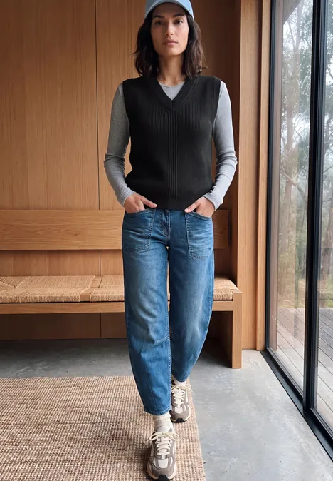 Pull sans manches uni à col en V Black Frau in blauer Mütze, grauem Shirt, schwarzer Weste und Jeans steht vor Holzbank am Fenster.