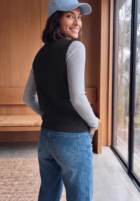 Pull sans manches uni à col en V Black Junge Frau in Jeans, grauem Shirt und schwarzer Weste, mit rückwärts getragener Cap, lächelnd am Fenster.