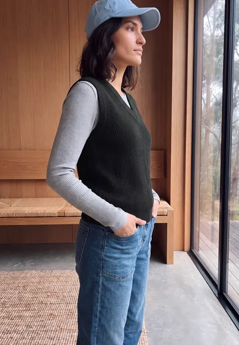 Pull sans manches uni à col en V Black Eine Frau mit Cap, grauem Shirt, schwarzer Weste und Jeans steht seitlich am Fenster.