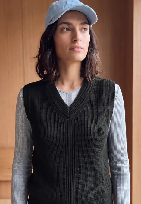 Pull sans manches uni à col en V Black Frau trägt schwarze Strickweste über grauem Langarmshirt und hellblauer Baseballkappe, vor hellem Hintergrund.