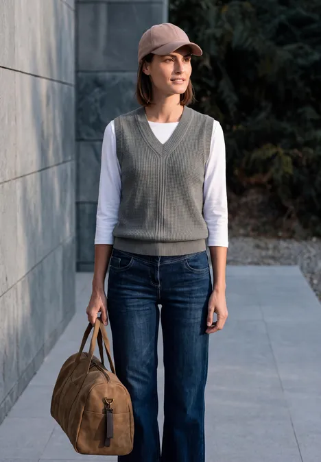 Femme en jean, gilet gris et casquette, tenant un sac de voyage marron devant un mur.