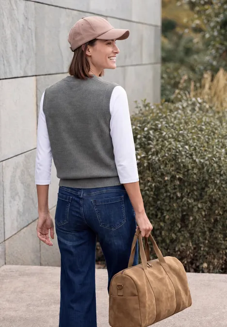 Femme de dos avec casquette, jean et gilet gris porte un sac de voyage marron le long d'un mur en pierre.