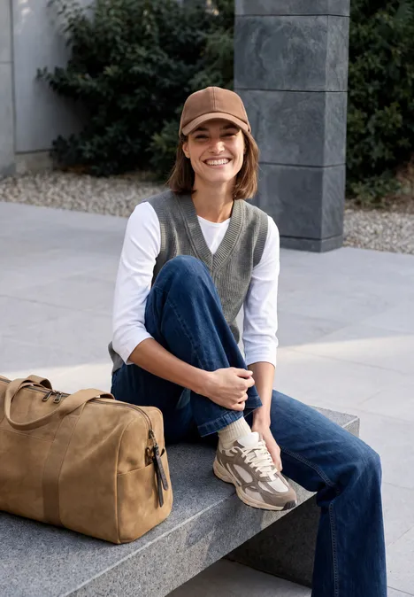 Femme en jean, t-shirt blanc, gilet vert et casquette, sourire, assise sur un banc en pierre avec sac.