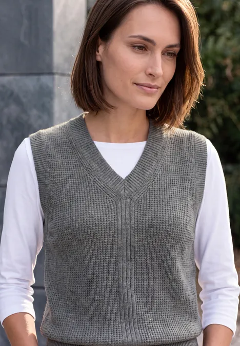 Femme aux cheveux bruns porte un gilet en maille gris sur un t-shirt blanc à manches longues devant un fond sobre