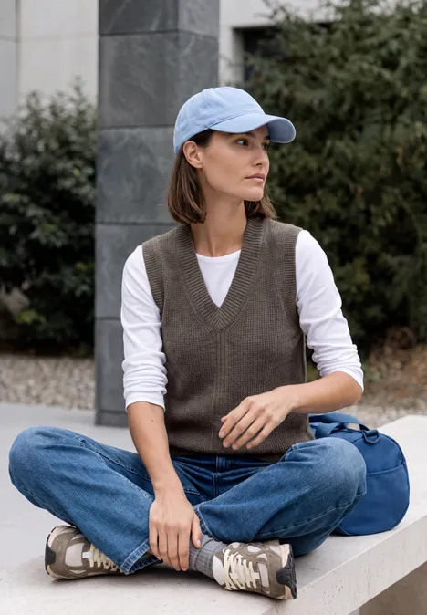 Femme portant casquette, jean et gilet, assise sur un muret en plein air, sac à côté.