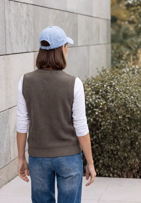 Personnage de dos, casquette blanche, tee-shirt à manches longues, gilet foncé, jeans, devant un mur et des buissons.