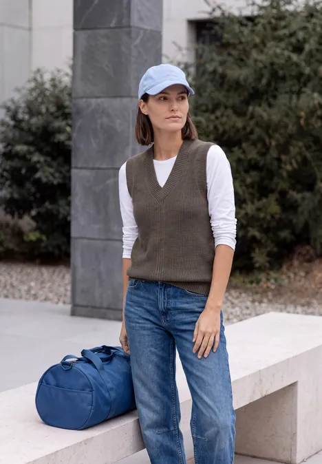 Femme en casquette, gilet tricoté, jean et sac bleu devant un décor urbain.