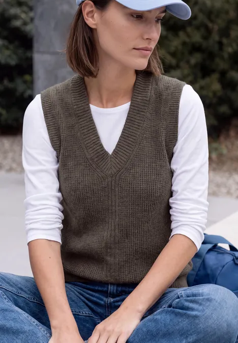 Une femme assise dehors porte un t-shirt à manches longues blanc, un gilet en maille marron, un jean et une casquette blanche.