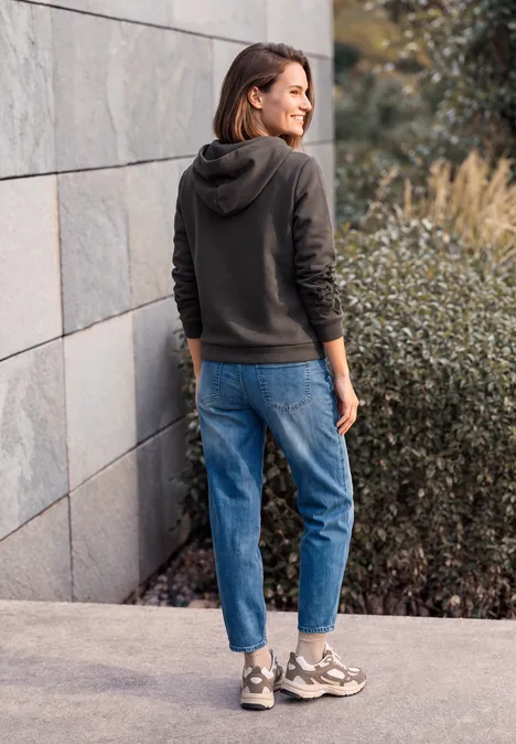 Frau in dunkler Kapuzenjacke und Jeans steht im Freien vor einer modernen Mauer.
