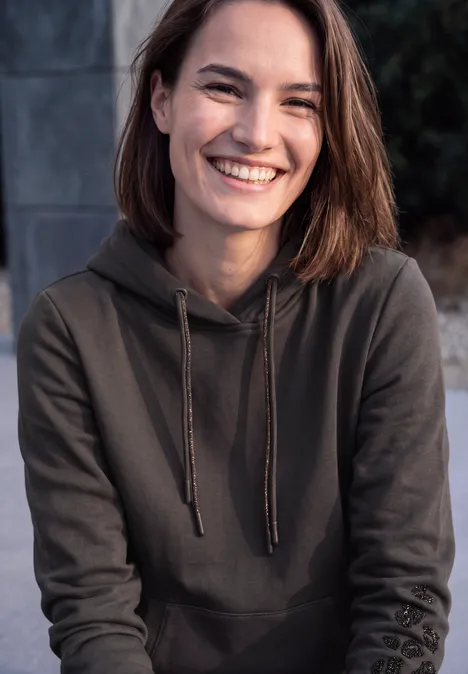 Eine junge Frau sitzt im Freien und lächelt, sie trägt ein dunkelgrünes Kapuzensweatshirt.