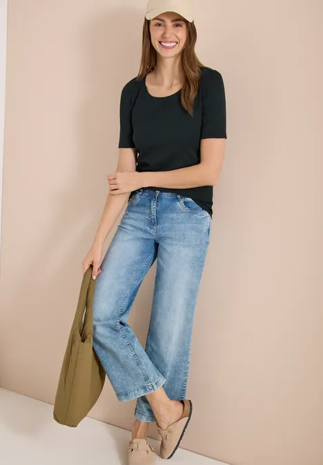 Vrouw in zwart T-shirt, blauwe jeans, beige schoenen en kaki tas, leunend tegen een beige muur.