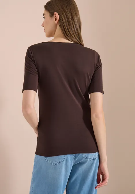 Femme de dos portant un top marron à manches courtes et un jean bleu clair sur fond neutre.