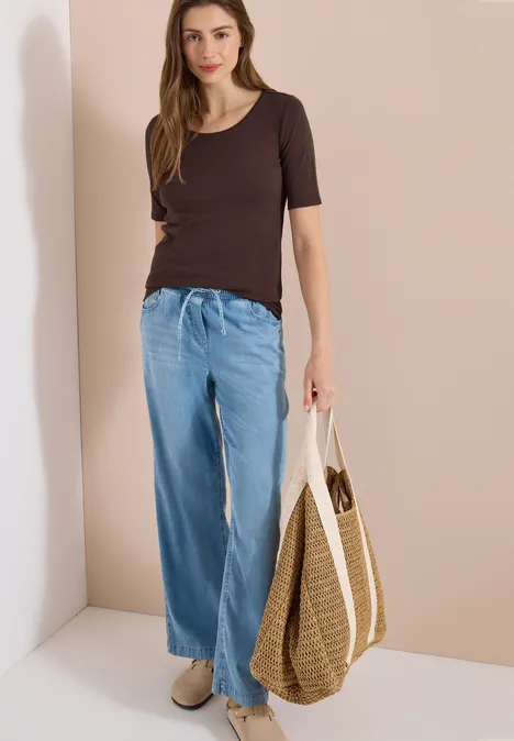 Femme en jean large, tee-shirt foncé, avec sac en paille et chaussures claires devant un mur beige.