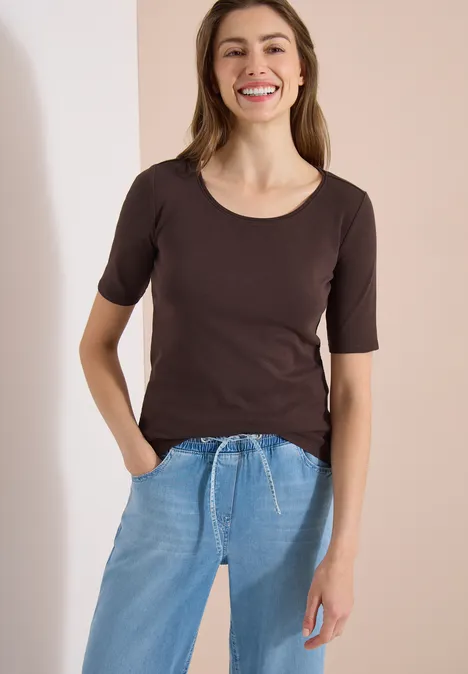 Femme porte un haut brun à manches courtes et un jean bleu clair à cordon, devant un fond clair.