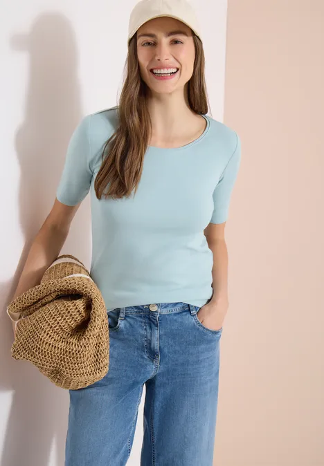 Femme en t-shirt bleu clair et jean tenant un sac en paille devant un fond clair.