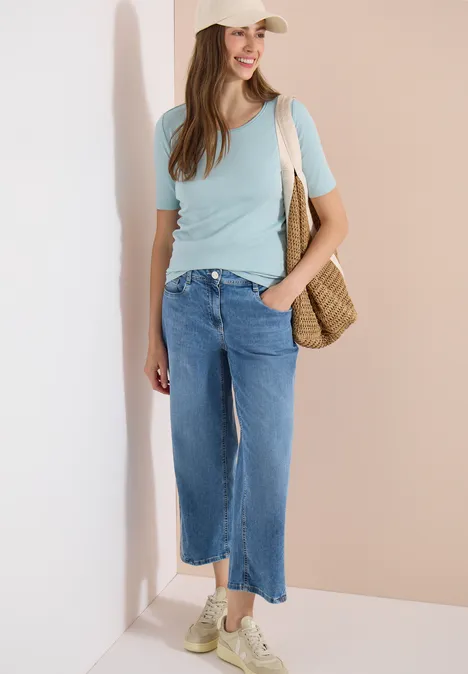 Femme en t-shirt bleu clair, jean ample et sac en paille, appuyée contre un mur beige.