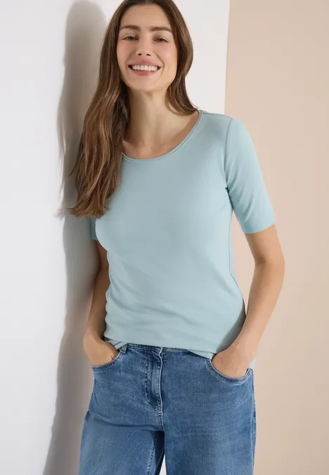 Femme appuyée contre un mur, portant un t-shirt bleu clair et un jean.