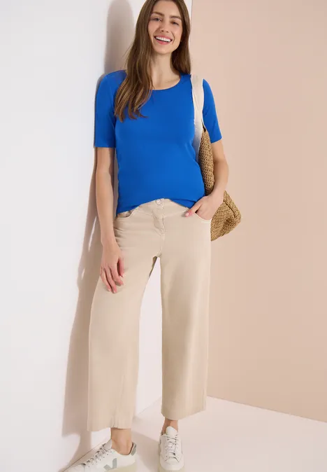 Femme appuyée contre un mur, haut bleu, pantalon ample beige, baskets blanches et sac en osier.