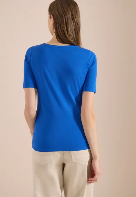 Femme vue de dos, porte un t-shirt bleu et un pantalon clair sur fond neutre.