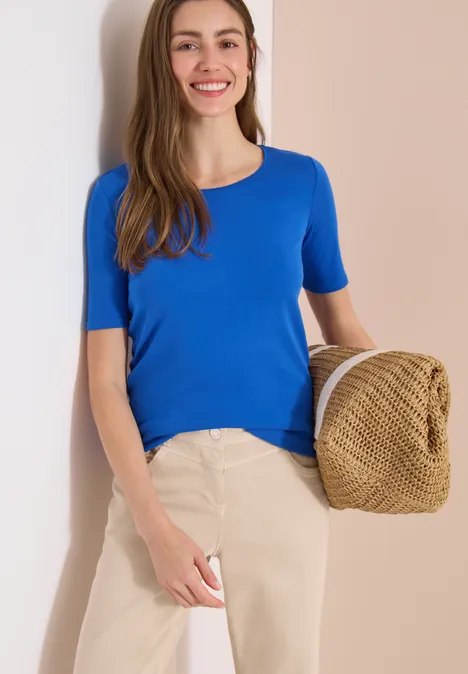 Femme penchée contre un mur clair, en t-shirt bleu, pantalon beige, tenant un chapeau en paille.