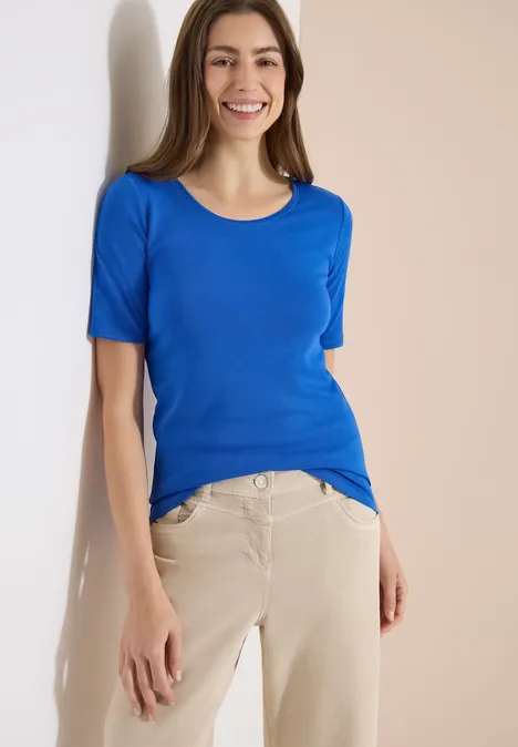 Une femme porte un t-shirt bleu et un pantalon beige devant un fond clair.