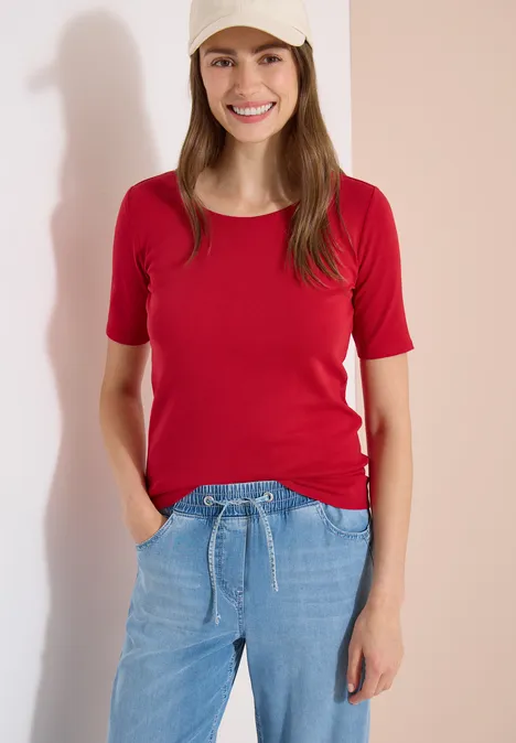 Femme devant un mur clair, vêtue d'un tee-shirt rouge et d'un jean bleu, mains dans les poches.