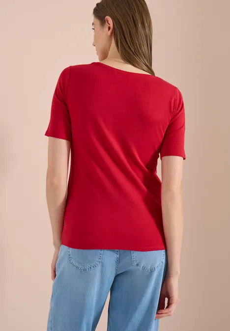 Femme de dos portant un t-shirt rouge et un jean bleu clair sur un fond neutre.