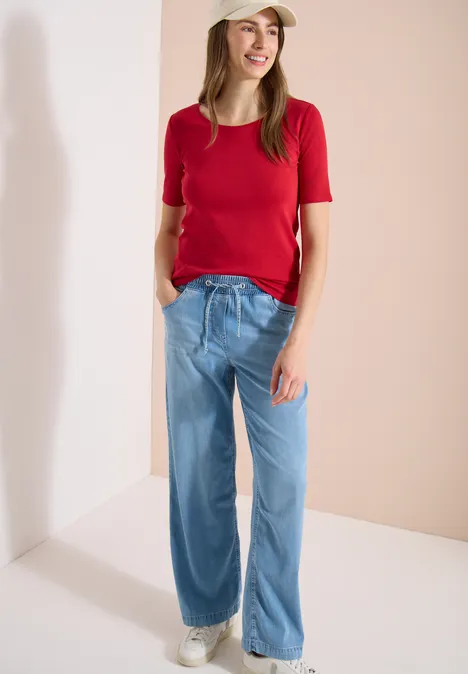 Femme en T-shirt rouge, jean ample bleu et baskets blanches devant un mur neutre.