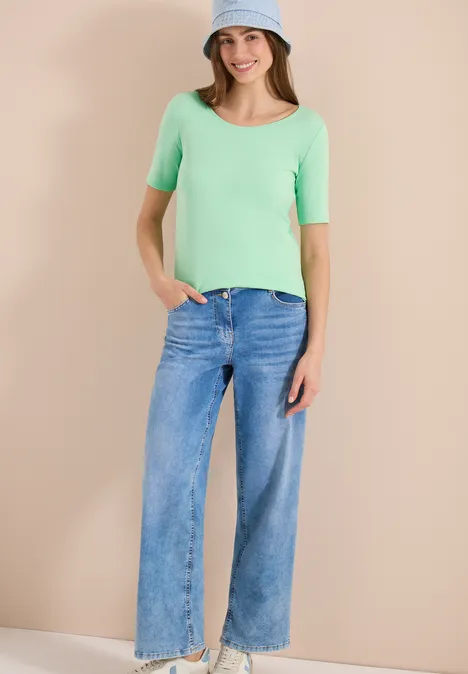 Frau steht vor beigem Hintergrund, trägt hellgrünes Oberteil mit halblangen Ärmeln und weite blaue Jeans.