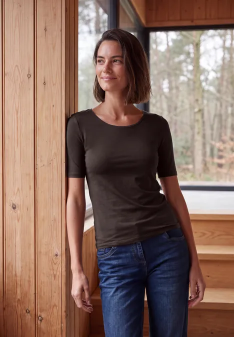 Frau lehnt entspannt an hölzernen Wand, Blick auf den Wald, sie trägt Jeans und ein dunkles Shirt.