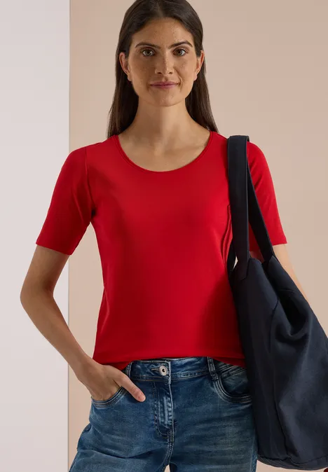 Femme en t-shirt rouge à manches courtes, jean et grand sac à l'épaule, fond clair.