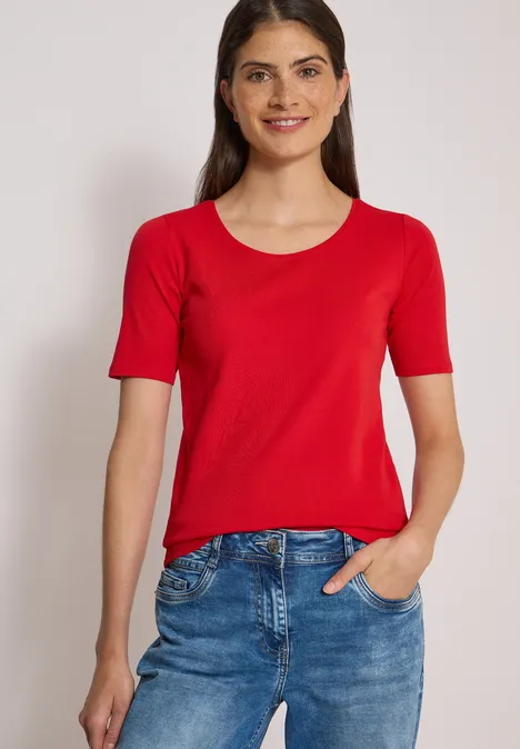 Femme en t-shirt rouge et jean bleu, souriante devant un fond clair.