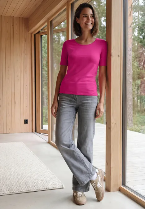 Basic Shirt met ronde hals in Unifarbe pink flash Vrouw lacht bij het raam, draagt een roze shirt, grijze jeans en lichte schoenen.