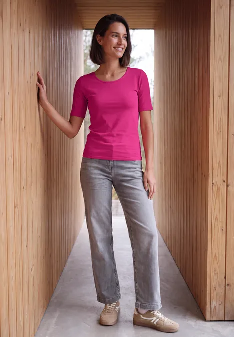 Basic Shirt met ronde hals in Unifarbe pink flash Een vrouw in een roze t-shirt, grijze jeans en beige sneakers leunt glimlachend in een houten gang.