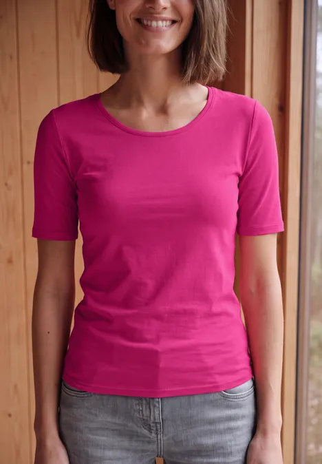 Basic Shirt met ronde hals in Unifarbe pink flash Een glimlachende vrouw in een roze T-shirt voor een houten achtergrond.