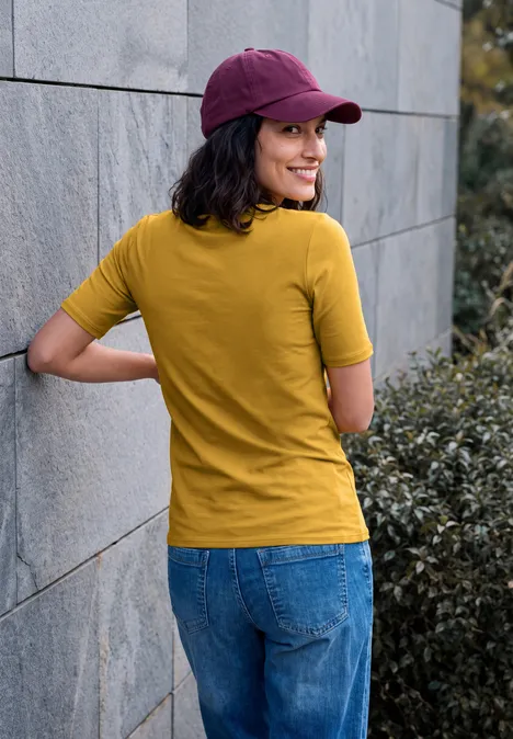 Frau im gelben T-Shirt, Jeans und lila Cap lehnt an einer Mauer und blickt lächelnd über die Schulter.