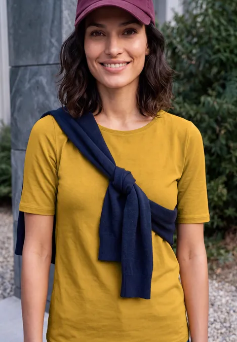 Eine lächelnde Frau in gelbem T-Shirt, mit dunkelblauem Pullover um die Schultern und rosa Cap.