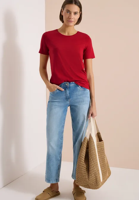 Frau trägt rotes T-Shirt, blaue Jeans und hält Strohtasche vor zweifarbigem Hintergrund.