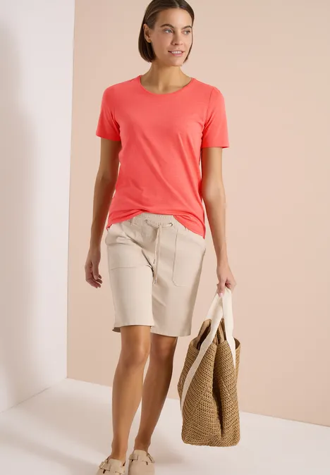Frau trägt korallenrotes T-Shirt, beige Bermudashorts und hält eine geflochtene Strandtasche.