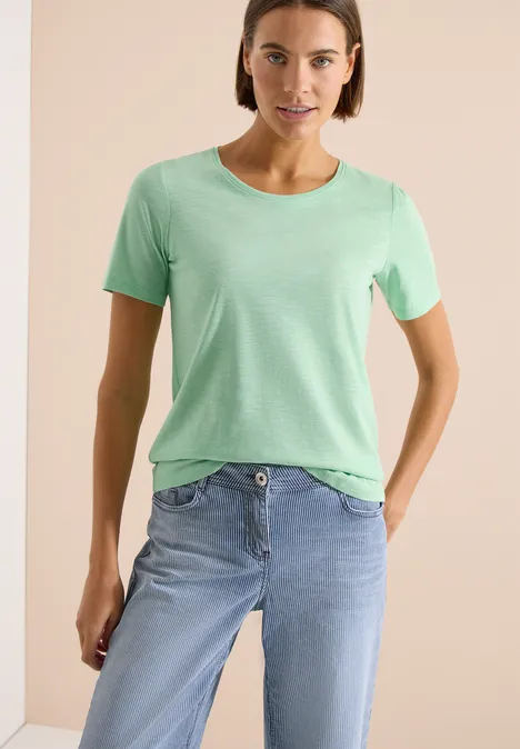 Vrouw in lichtgroen T-shirt en lichtblauwe spijkerbroek voor een neutrale achtergrond.