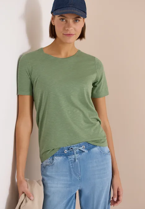 Person in grünem T-Shirt und hellblauen Jeans lehnt an einer Wand.