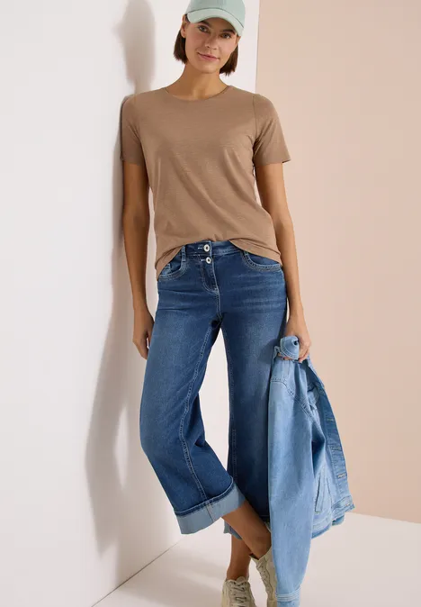 Basic Shirt met ronde hals creamy mocha Vrouw geleund tegen muur, bruine t-shirt, blauwe jeans, spijkerjasje in hand.