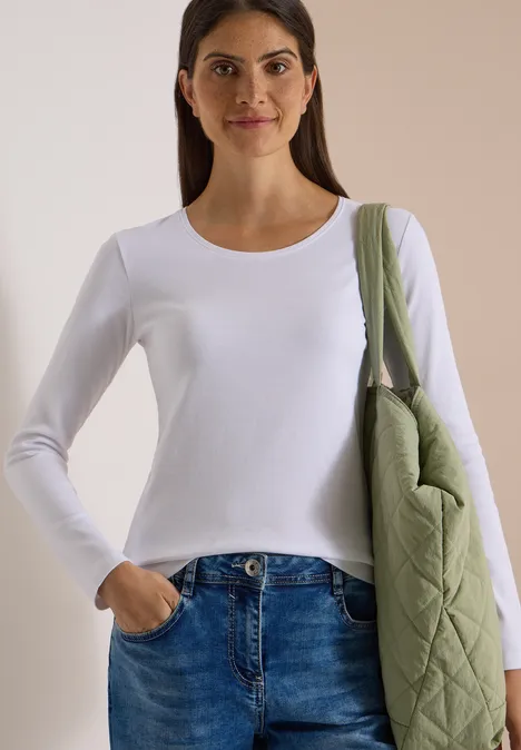 Basic shirt met lange mouwen en ronde hals White Eine Frau in weißem Langarmshirt und Jeans, mit grünem Stepp-Schulterbeutel vor neutralem Hintergrund.