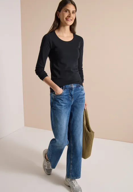 Basic shirt met lange mouwen en ronde hals Black Frau in schwarzem Langarmshirt, blauen Jeans und Sneakern hält eine grüne Tasche und lächelt.