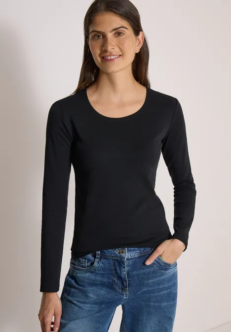 Basic shirt met lange mouwen en ronde hals Black Frau mit schwarzem Langarmshirt und blauen Jeans lächelt vor hellem Hintergrund.