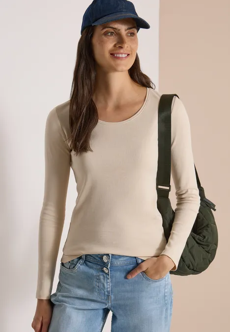 Basic shirt met lange mouwen en ronde hals pearl beige Frau lächelt, trägt beiges Langarmshirt, Jeans, blaue Kappe und grüne Tasche über der Schulter.