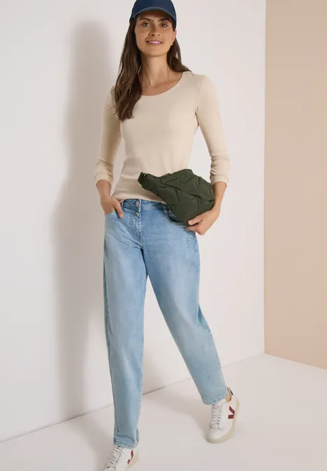 Basic shirt met lange mouwen en ronde hals pearl beige Frau in Jeans, weißem Langarmshirt, Cap und Umhängetasche lehnt an heller Wand.