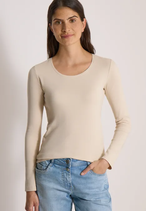 Basic shirt met lange mouwen en ronde hals pearl beige Junge Frau in beigefarbenem Langarmshirt und blauen Jeans steht lächelnd mit einer Hand in der Hosentasche.