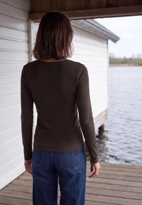 Eine Person in langärmligem dunklem Shirt und Jeans steht am Steg vor einem Bootshaus und blickt aufs Wasser.