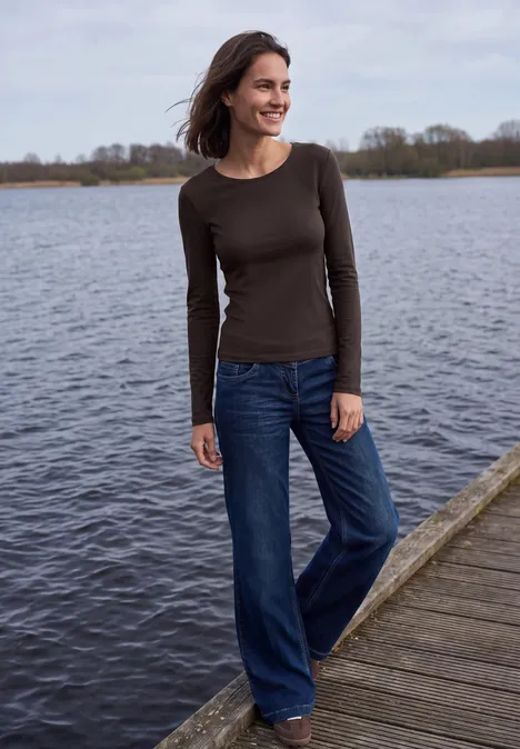 Frau in braunem Langarmshirt und weitem Jeans steht lächelnd auf einem Steg am See